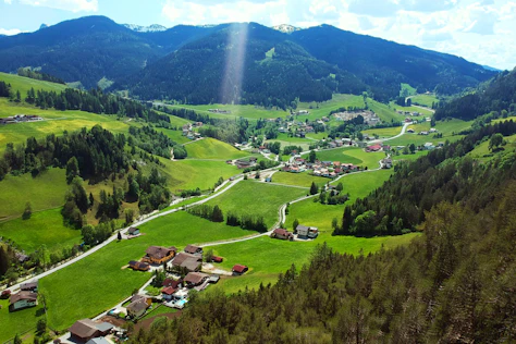 Erhöhter Blick auf ein idyllisches Bergtal mit verstreuten Häusern, grünen Wiesen und bewaldeten Hängen. Kleine Straßen schlängeln sich durch die Landschaft, während im Hintergrund sanfte Berge unter hellem Sonnenlicht aufragen.