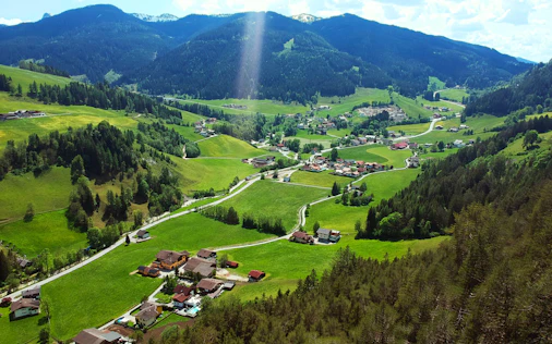 Erhöhter Blick auf ein idyllisches Bergtal mit verstreuten Häusern, grünen Wiesen und bewaldeten Hängen. Kleine Straßen schlängeln sich durch die Landschaft, während im Hintergrund sanfte Berge unter hellem Sonnenlicht aufragen.