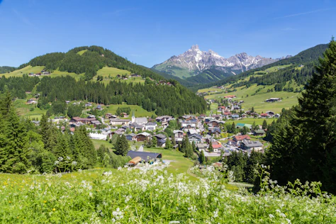 Erhöhter Blick auf ein alpines Dorf mit Kirche im Tal, umgeben von grünen Wiesen und dichten Wäldern. Im Hintergrund ragen markante, teils schneebedeckte Berggipfel unter einem klaren blauen Himmel auf.