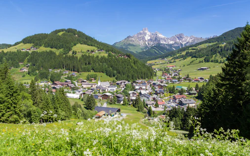 Erhöhter Blick auf ein alpines Dorf mit Kirche im Tal, umgeben von grünen Wiesen und dichten Wäldern. Im Hintergrund ragen markante, teils schneebedeckte Berggipfel unter einem klaren blauen Himmel auf.