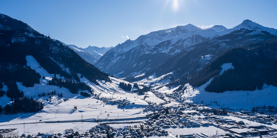 Panoramablick auf ein verschneites Tal mit einer Ortschaft im Vordergrund, umgeben von steilen, schneebedeckten Berghängen. Die Wintersonne steht hoch am Himmel und taucht die alpine Landschaft in klares, kaltes Licht.