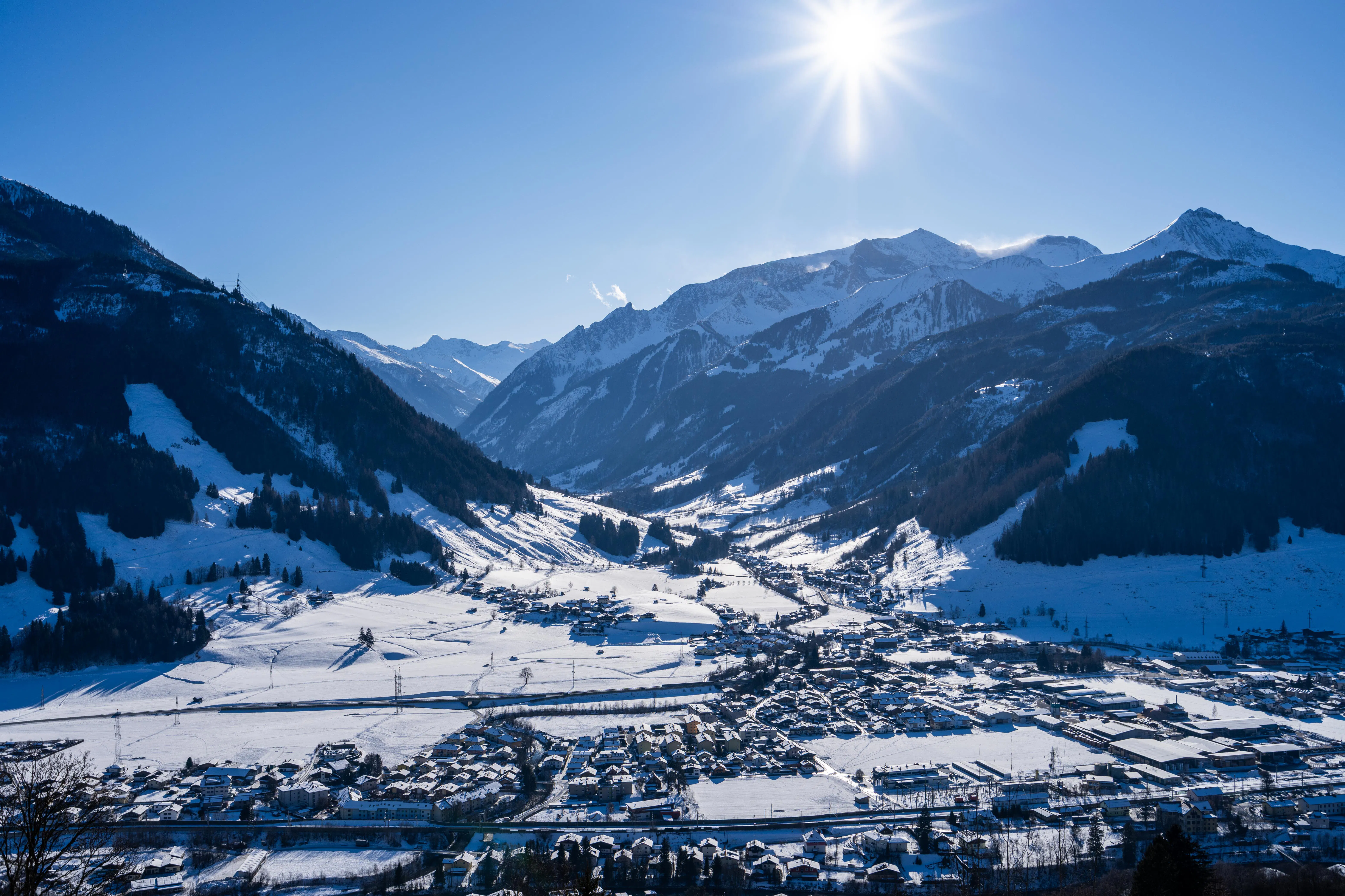 Panoramic view of a snow-covered valley with a village in the foreground, surrounded by steep, snow-covered mountain slopes. The winter sun shines high in the sky, casting clear, crisp light over the alpine landscape.