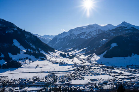 Panoramablick auf ein verschneites Tal mit einer Ortschaft im Vordergrund, umgeben von steilen, schneebedeckten Berghängen. Die Wintersonne steht hoch am Himmel und taucht die alpine Landschaft in klares, kaltes Licht.