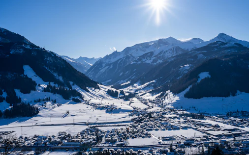 Panoramablick auf ein verschneites Tal mit einer Ortschaft im Vordergrund, umgeben von steilen, schneebedeckten Berghängen. Die Wintersonne steht hoch am Himmel und taucht die alpine Landschaft in klares, kaltes Licht.