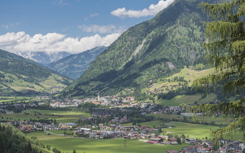 A wide alpine valley surrounded by steep green mountains under a partly cloudy sky. In the center, a small town with clustered houses lies along the valley floor, bordered by fields and meadows.