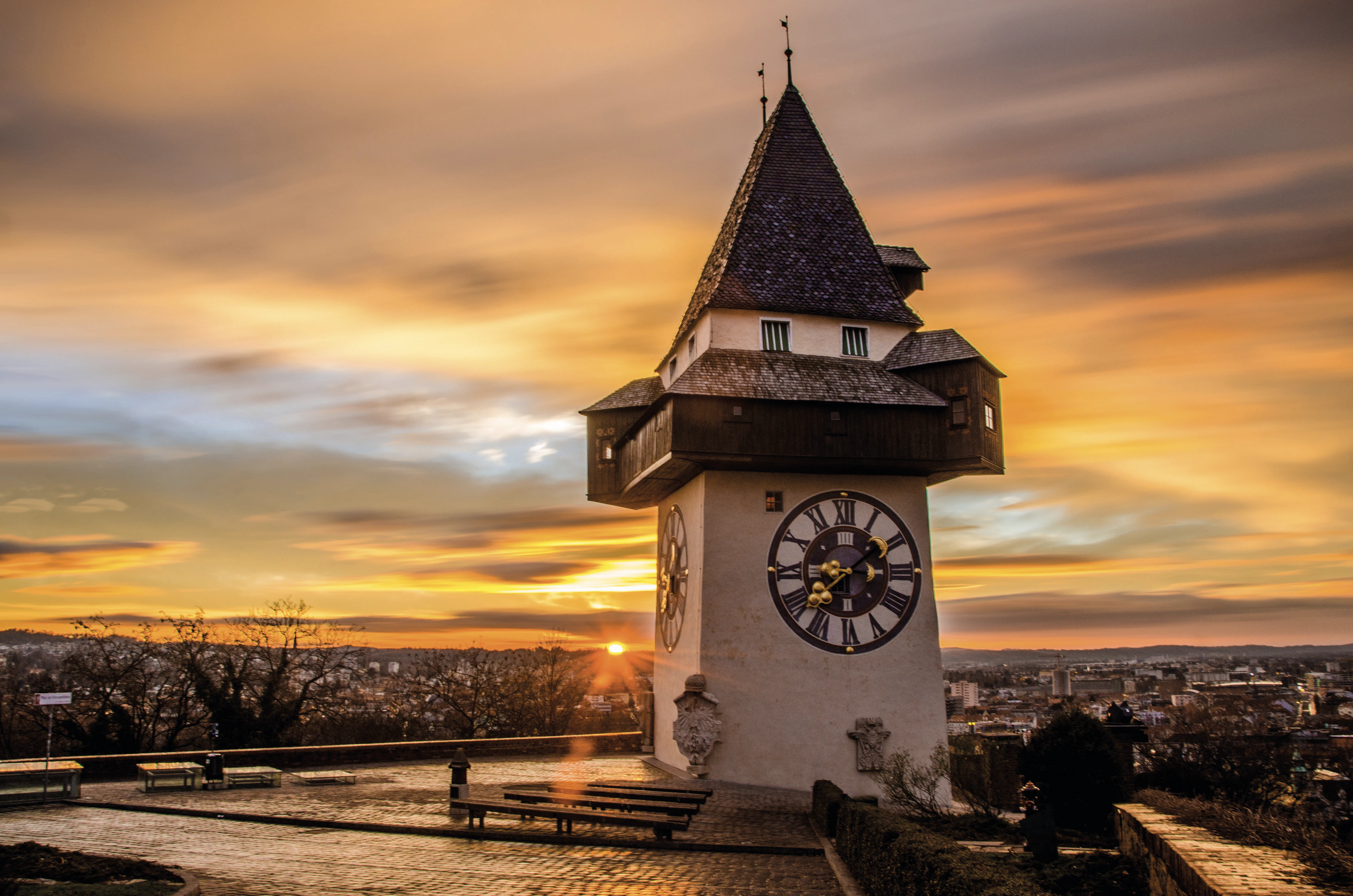 Historischer Uhrturm mit Spitzdach bei Sonnenuntergang, mit Blick über die Dächer von Bregenz und den Bodensee im Hintergrund.