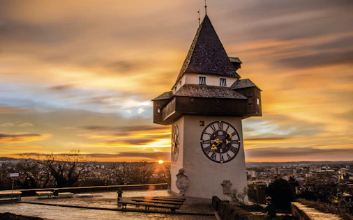 Historischer Uhrturm mit Spitzdach bei Sonnenuntergang, mit Blick über die Dächer von Bregenz und den Bodensee im Hintergrund.