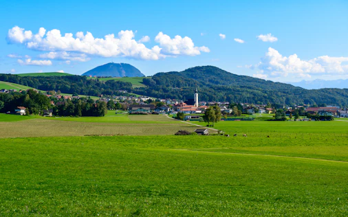 Ein weites grünes Feld erstreckt sich in Richtung eines kleinen Ortes, dahinter erheben sich Berge unter einem strahlend blauen Himmel mit vereinzelten Wolken.