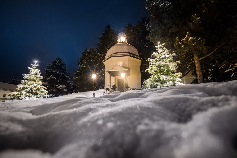 A small illuminated chapel stands quietly in a snowy landscape at night, surrounded by trees and soft, glowing lights.
