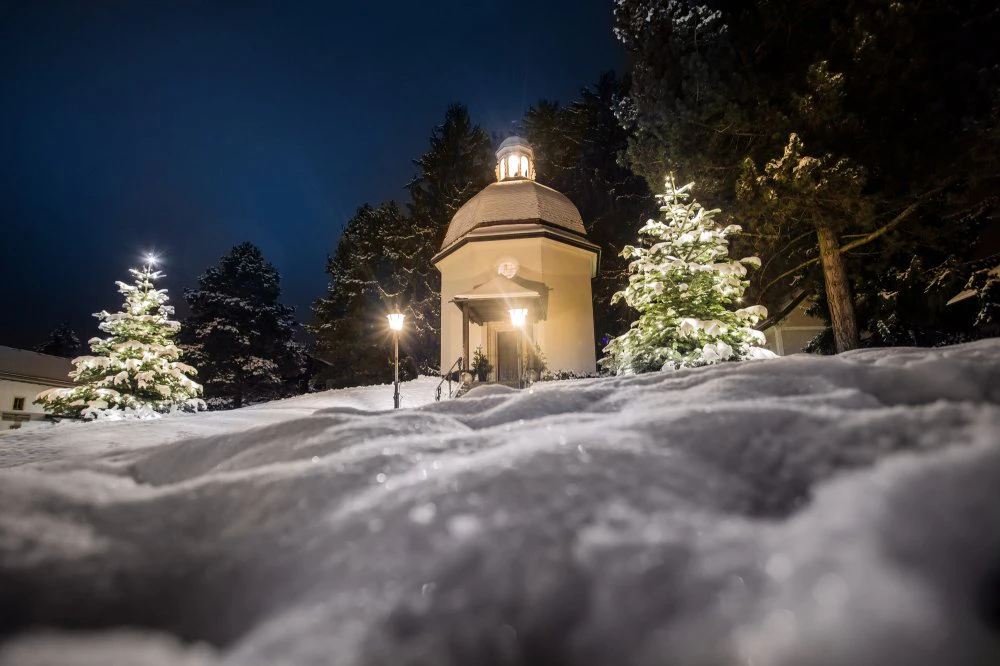 A small illuminated chapel stands quietly in a snowy landscape at night, surrounded by trees and soft, glowing lights.