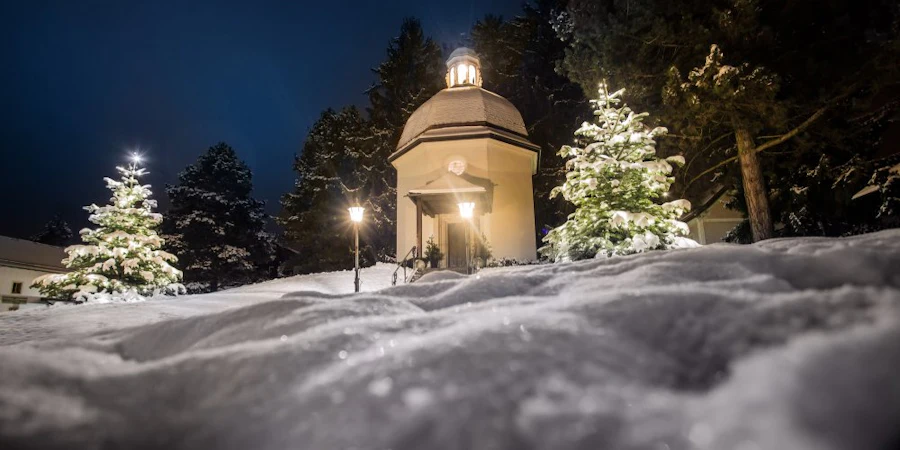 A small illuminated chapel stands quietly in a snowy landscape at night, surrounded by trees and soft, glowing lights.