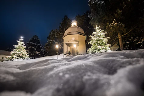 A small illuminated chapel stands quietly in a snowy landscape at night, surrounded by trees and soft, glowing lights.
