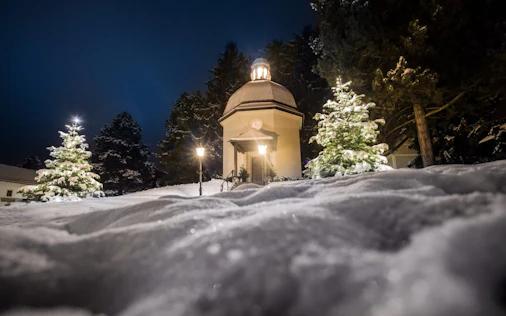 Eine kleine, beleuchtete Kapelle steht ruhig in einer verschneiten Landschaft bei Nacht, umgeben von Bäumen und sanftem Licht.