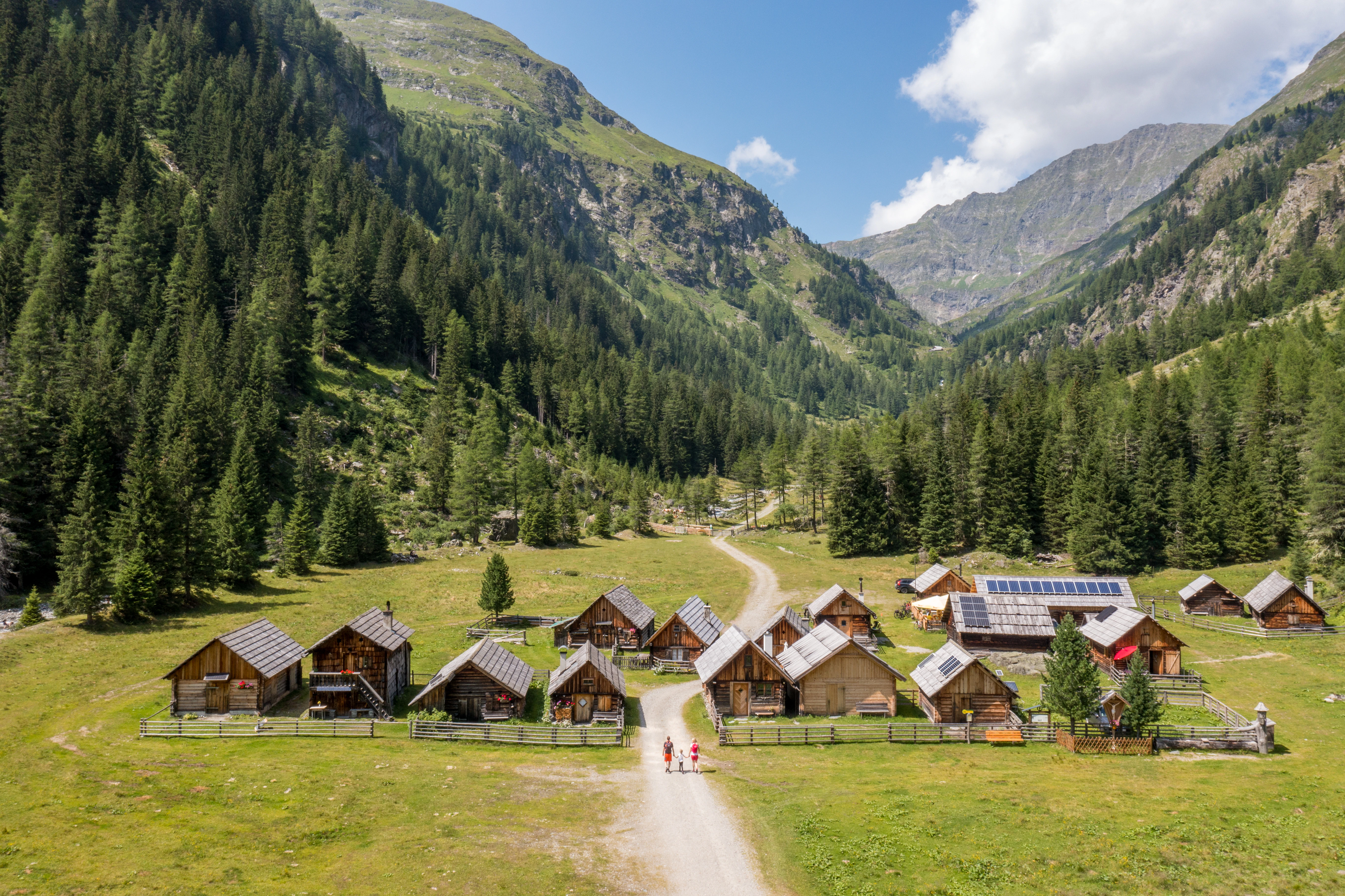 Wide mountain landscape with green meadows, in the center a small village with several wooden houses and a church, surrounded by high, forested mountains under a clear sky.