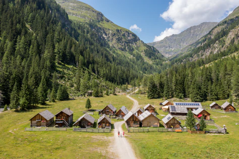 Wide mountain landscape with green meadows, in the center a small village with several wooden houses and a church, surrounded by high, forested mountains under a clear sky.