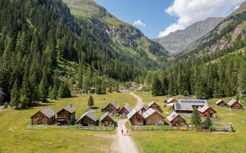 Weite Berglandschaft mit grünen Wiesen, in der Mitte ein kleines Dorf mit mehreren Holzhäusern und einer Kirche, umgeben von hohen, bewaldeten Bergen unter klarem Himmel. übersetze es auf englisch
