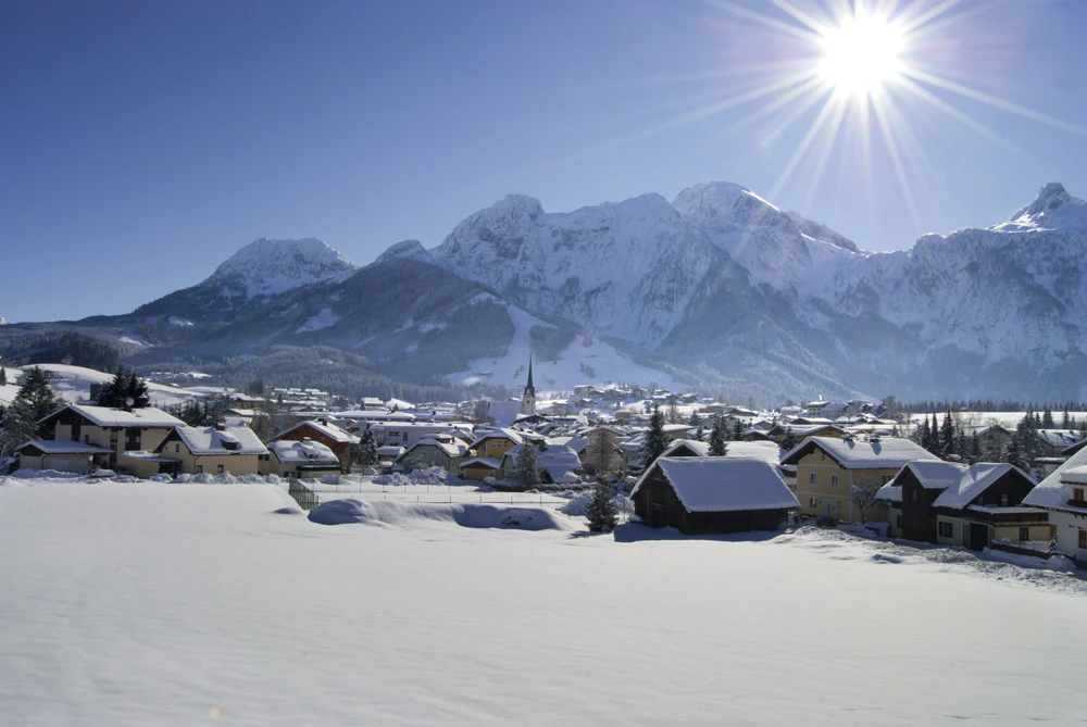 Winterliche Berglandschaft mit verschneitem Dorf im Tal, umgeben von hohen Bergen unter strahlendem Sonnenschein.