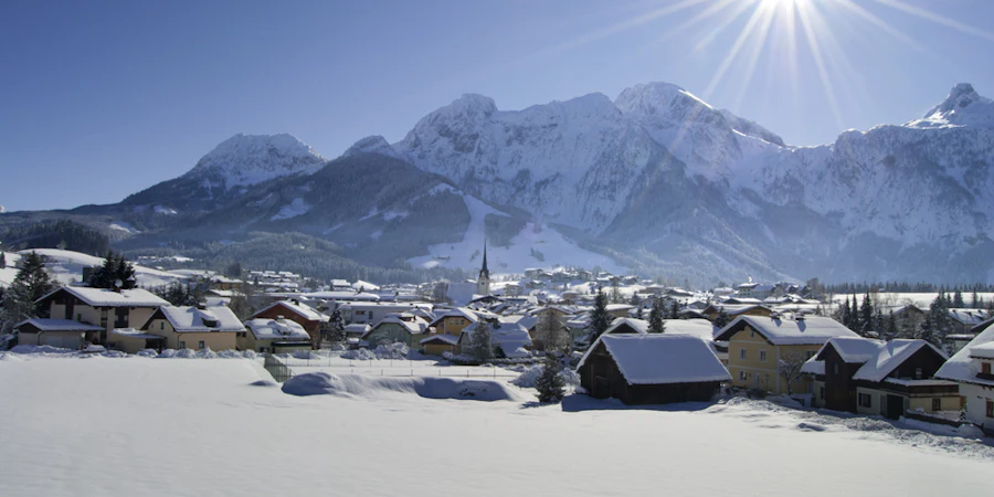 Winterliche Berglandschaft mit verschneitem Dorf im Tal, umgeben von hohen Bergen unter strahlendem Sonnenschein.