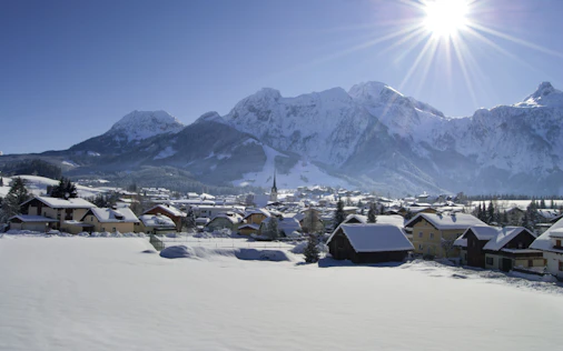 Winterliche Berglandschaft mit verschneitem Dorf im Tal, umgeben von hohen Bergen unter strahlendem Sonnenschein.