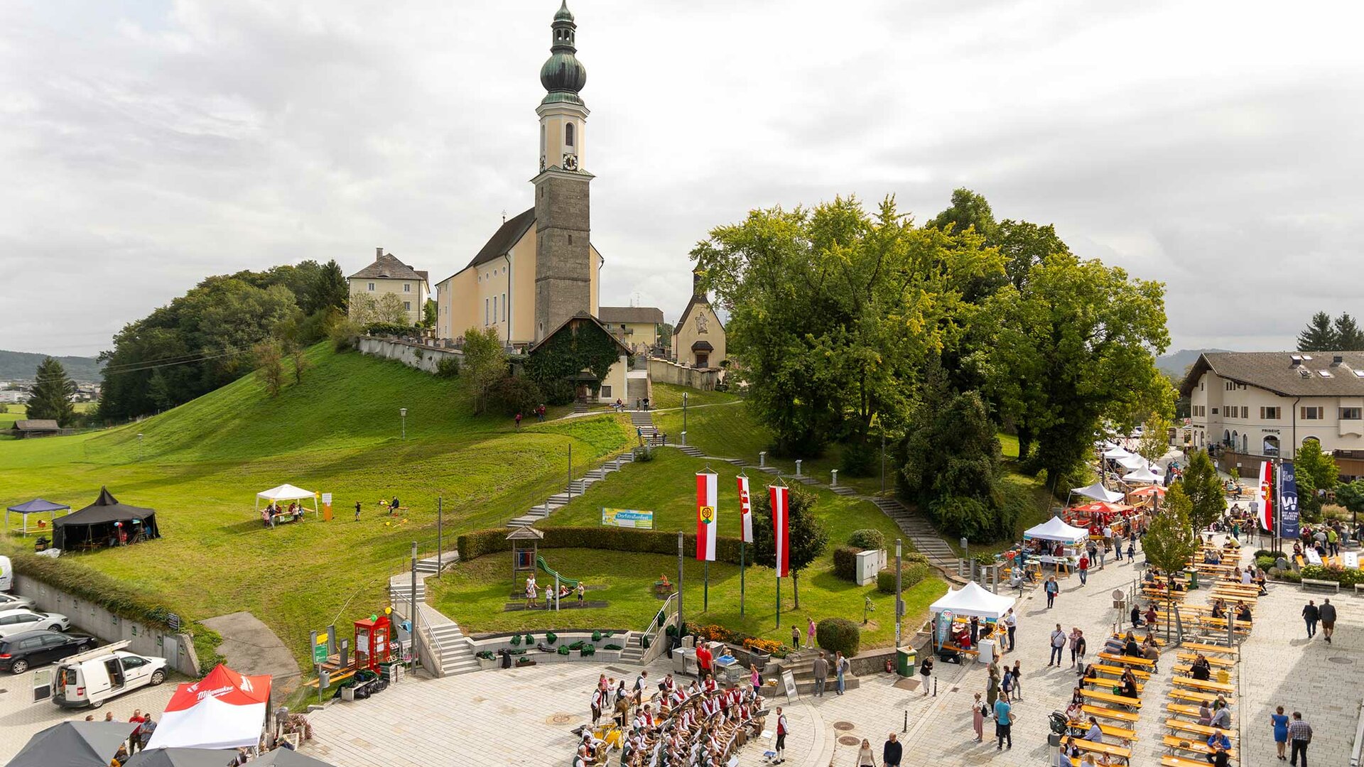 Luftaufnahme einer Kirche auf einem Hügel in Bergheim, umgeben von grünen Wiesen und einem kleinen Ortskern mit Häusern und Straßen.