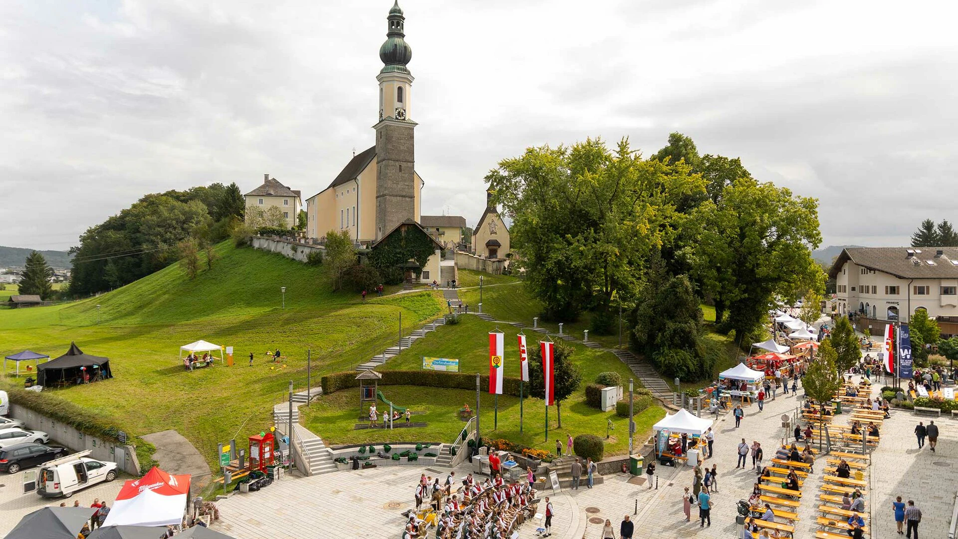 Luftaufnahme einer Kirche auf einem Hügel in Bergheim, umgeben von grünen Wiesen und einem kleinen Ortskern mit Häusern und Straßen.
