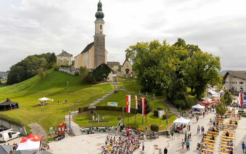 Luftaufnahme einer Kirche auf einem Hügel in Bergheim, umgeben von grünen Wiesen und einem kleinen Ortskern mit Häusern und Straßen.