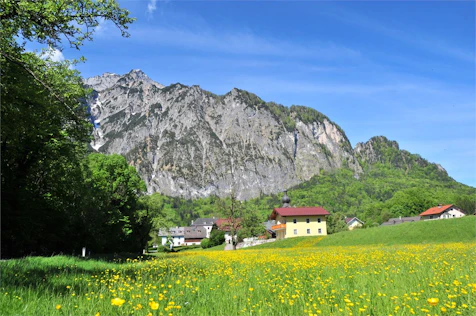 The image shows a sunny mountain landscape with a blooming meadow in the foreground and a few houses at the foot of a striking rock formation. The combination of nature, tranquility, and alpine scenery conveys a high quality of life and relaxation.