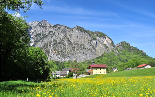 Das Bild zeigt eine sonnige Berglandschaft mit blühender Wiese im Vordergrund und einzelnen Häusern am Fuße eines markanten Felsmassivs. Die Kombination aus Natur, Ruhe und alpiner Kulisse vermittelt hohe Lebensqualität und Erholung.