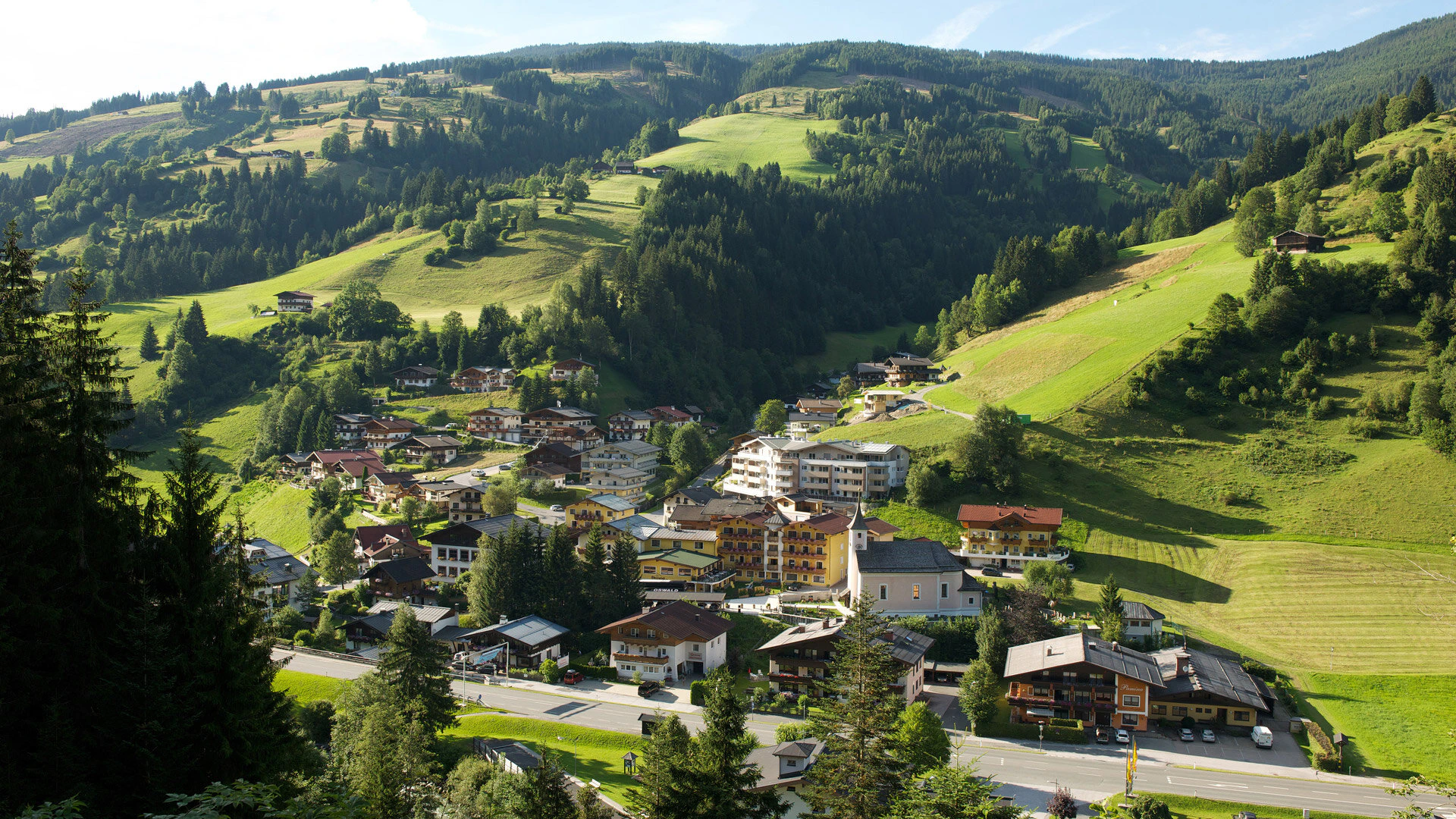The image shows an idyllic alpine village nestled in gentle green hills. The scattered houses and open landscape convey a sense of tranquility connection to nature and an authentic rural lifestyle.
