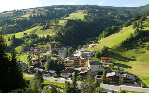 Das Bild zeigt ein idyllisches Alpendorf eingebettet in sanfte, grüne Hügel. Die verstreuten Häuser und die offene Landschaft vermitteln Ruhe, Naturverbundenheit und ein authentisches Lebensgefühl im ländlichen Raum.