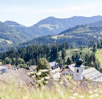 Ein ruhiger Ausblick auf eine grüne Hügellandschaft mit vereinzelten Häusern, eingebettet in die Natur. Im Hintergrund erheben sich sanfte Berge, die dem Bild eine entspannte, alpine Atmosphäre verleihen.
