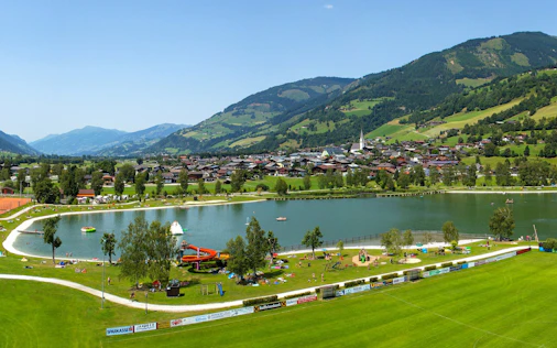 Ein idyllischer See eingebettet in eine grüne Landschaft mit Blick auf sanfte Hügel und Berge, ergänzt durch gepflegte Wege und eine ruhige, einladende Atmosphäre.