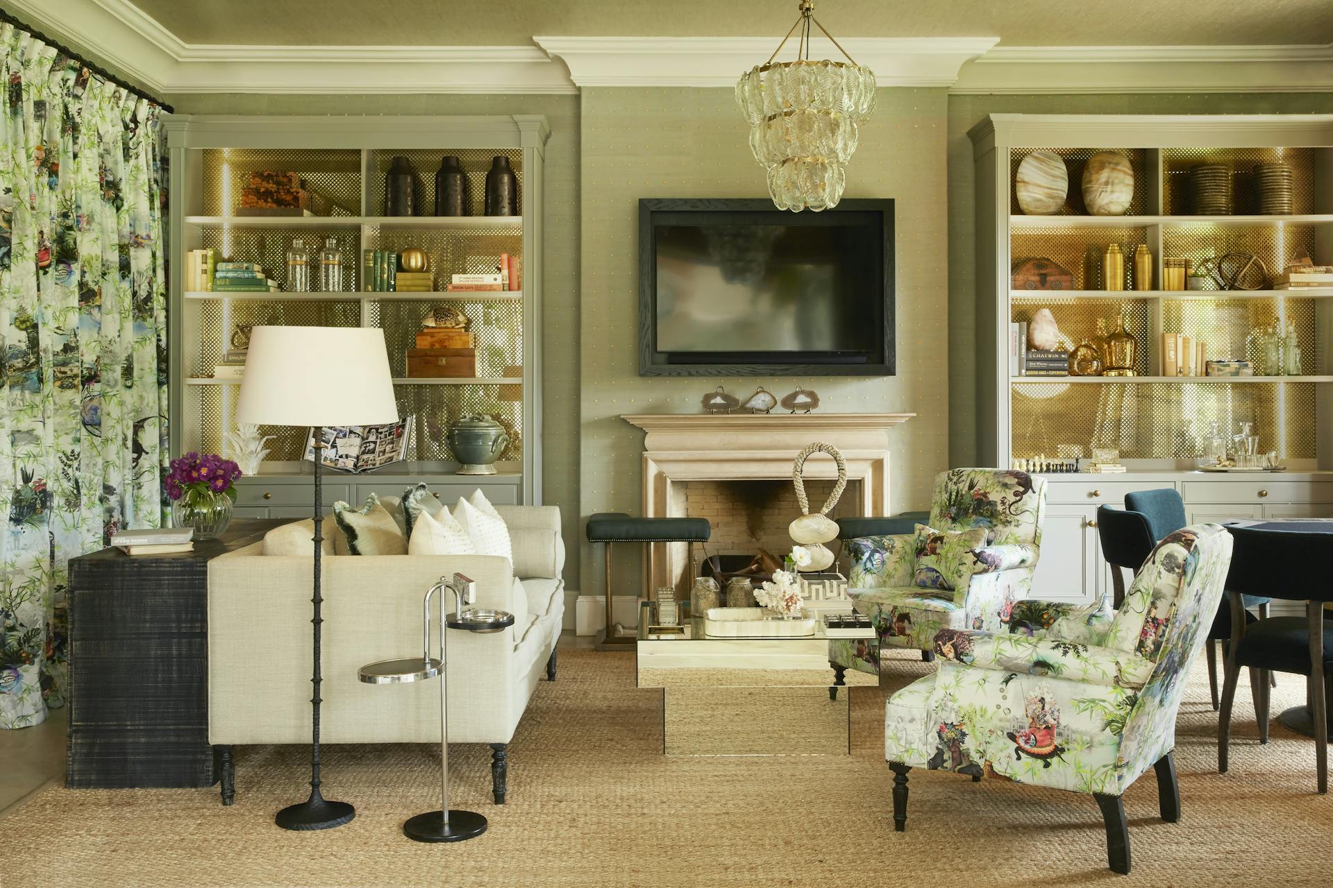 A sitting room features a square, mirrored coffee table, cream-coloured sofa, armchairs upholstered in ornate fabric, and two bookshelves and bookshelves holding vintage books alongside decorative items.