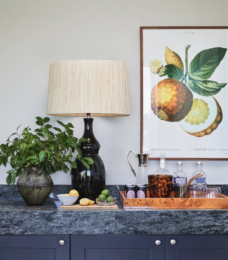 A lamp, a tray of alcohol, a plant is seen on a console table with fruit artwork on the wall just behind.