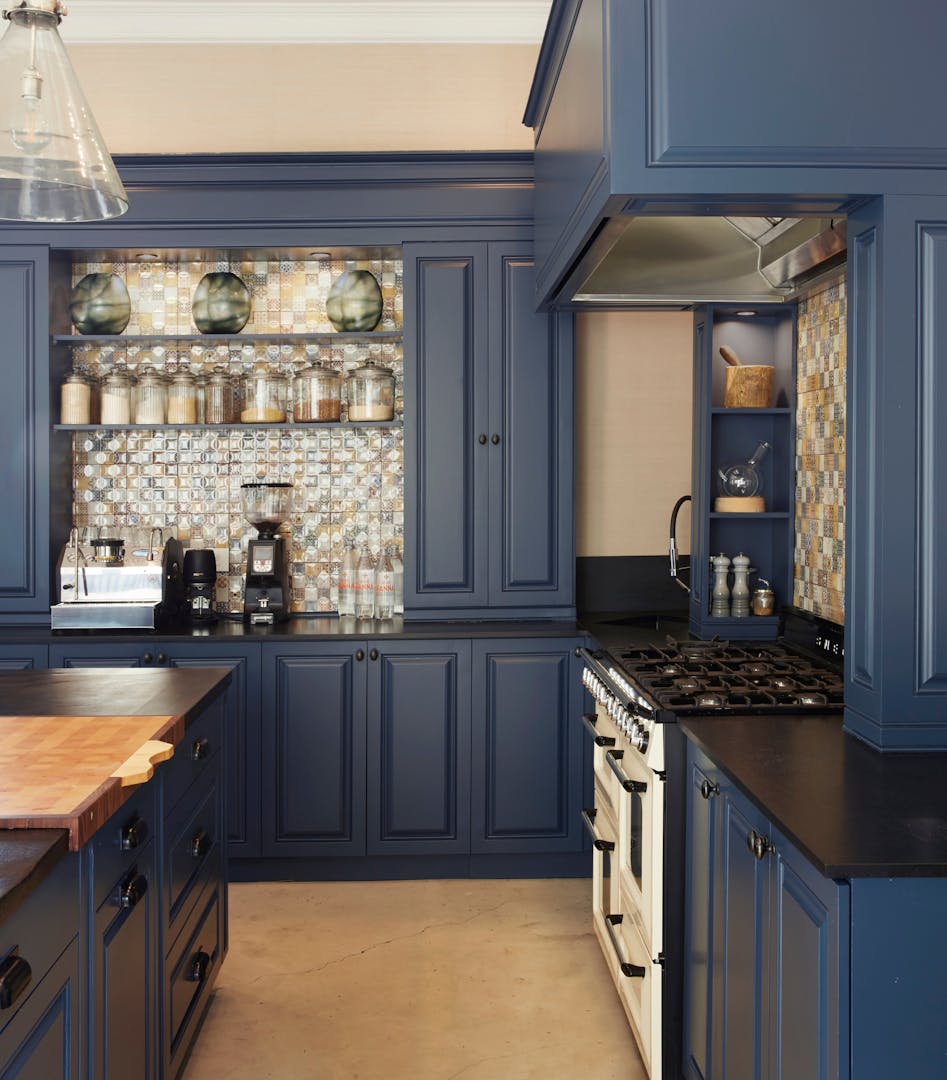 A section of a kitchen displays dark blue cabinets, black countertops and a wooden work surface. Sections of the wall are visible and feature patterned tiles, in front of which are a coffee machine, coffee bean grinder, and jars of spices and dry goods.