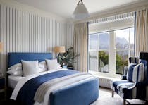 A contemporary bedroom with blue and white striped wallpaper, a navy blue bed layered in blue, white and grey bedding and an armchair in the corner next to a window with a view of trees.