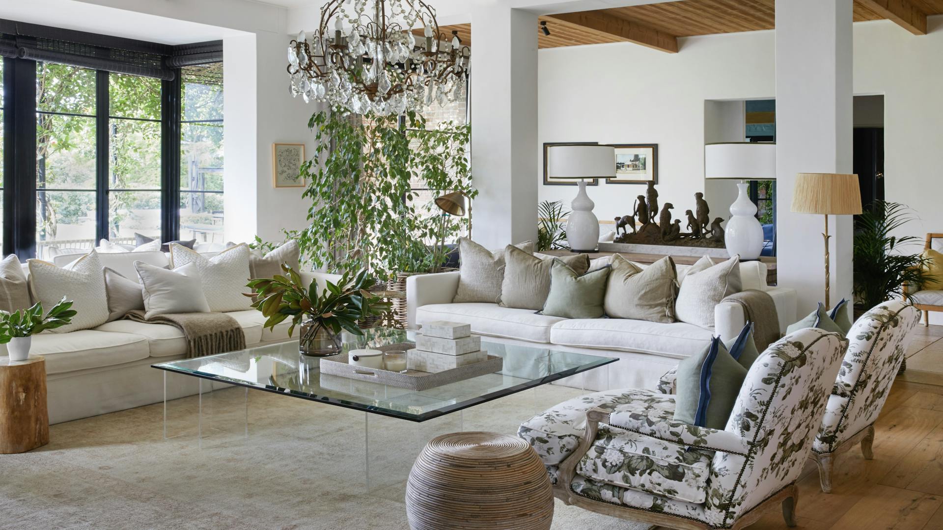 A sitting area features two couches, upholstered chairs, and a glass coffee table holding a tray of square objects and a glass vase of greenery. A leafy plant, a pair of white lamps, and a sculpture of a group of meerkats are visible in the background. A chandelier hangs above