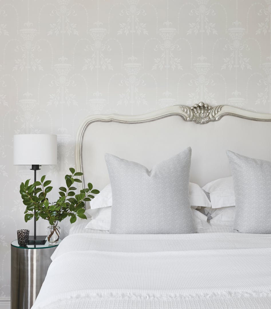 Close-up of a bed with white linen and circular, steel side table with a modern lamp on top of it.