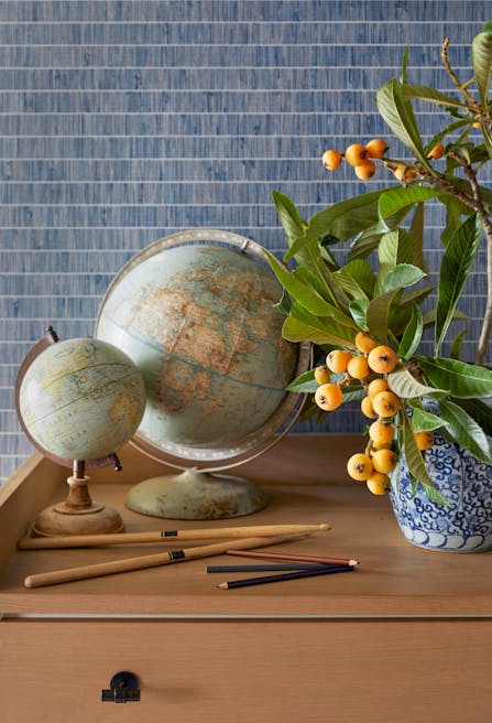 Drumsticks, pencils and two globes of differing sizes sit on a wooden surface beside a blue-patterned ceramic vase holding greenery and fruit that may be loquats.