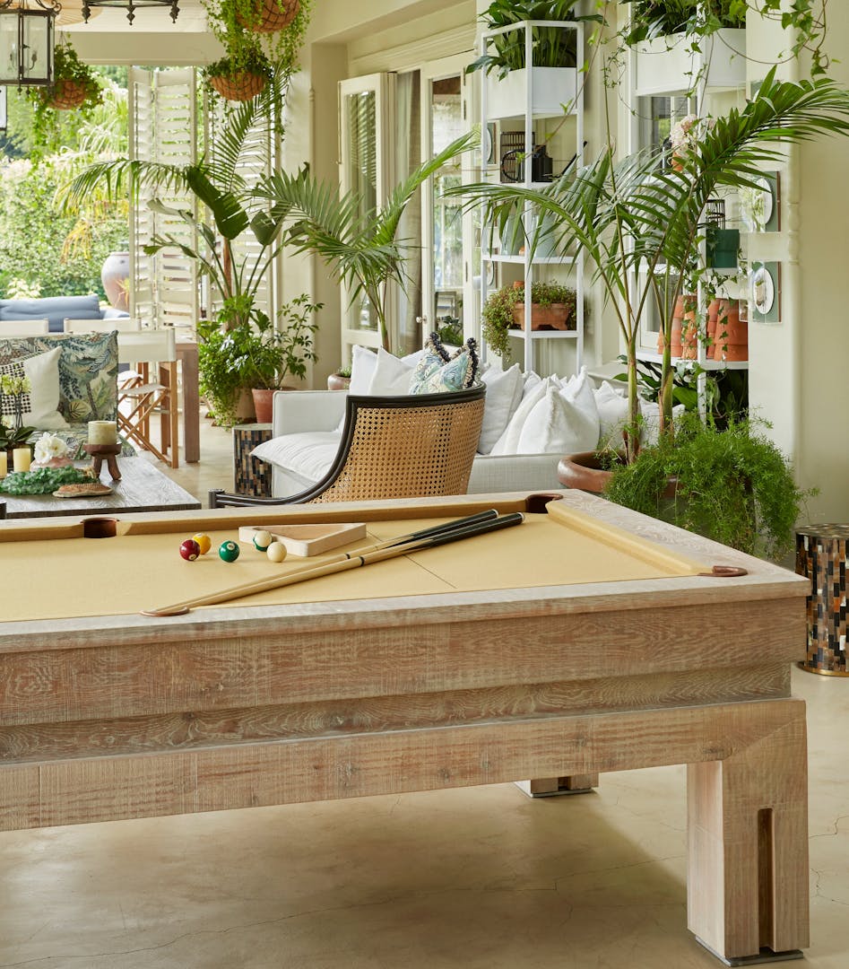 A wooden pool table in the foreground with an outdoor entertainment area that features rattan armchairs and linen sofas in the background.
