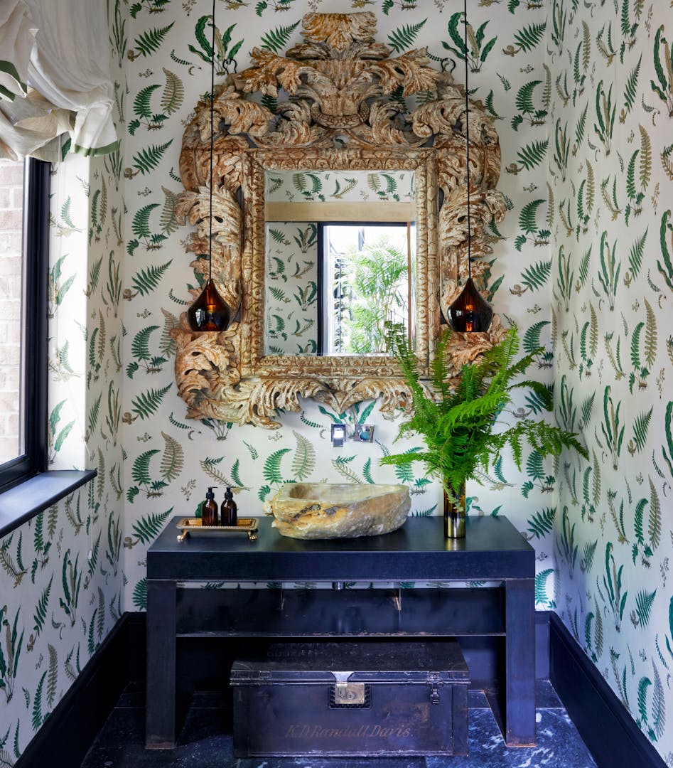 A sink hollowed out of a large rock sits on a deep blue vanity, below an ornately carved wood-framed mirror. The wallpaper is patterned with illustrated ferns and two brown glass pendant links hang in front of the mirror