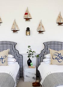 A marine-inspired bedroom with two single beds that both feature blue and white checkered headboards. Artwork of ships hang on the wall with a side table in between the beds.