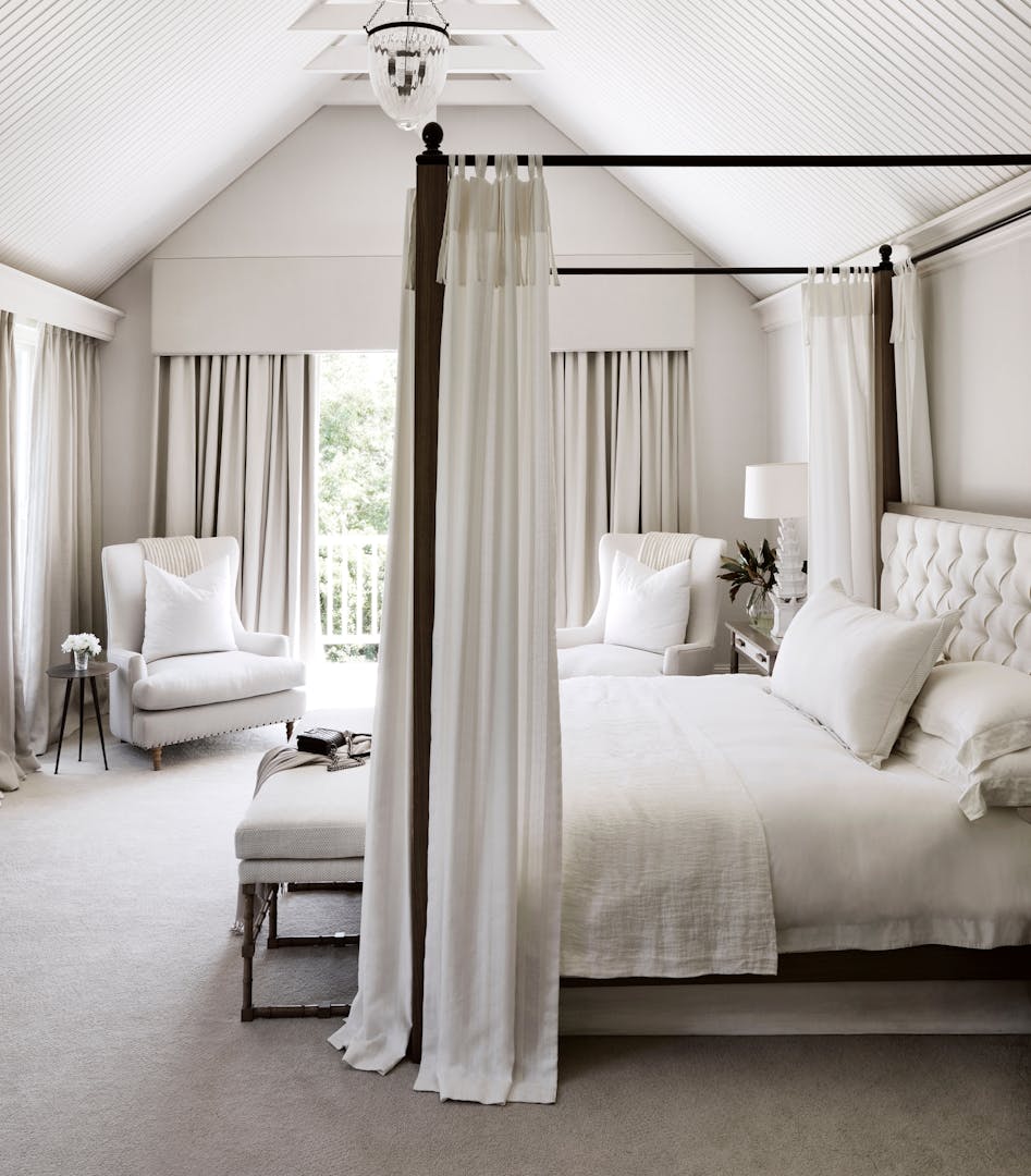 A four-poster bed, upholstered chairs, bedroom bench and side table are seen in a bedroom with an A-frame ceiling, furnished in white, light neutrals and dark wood.