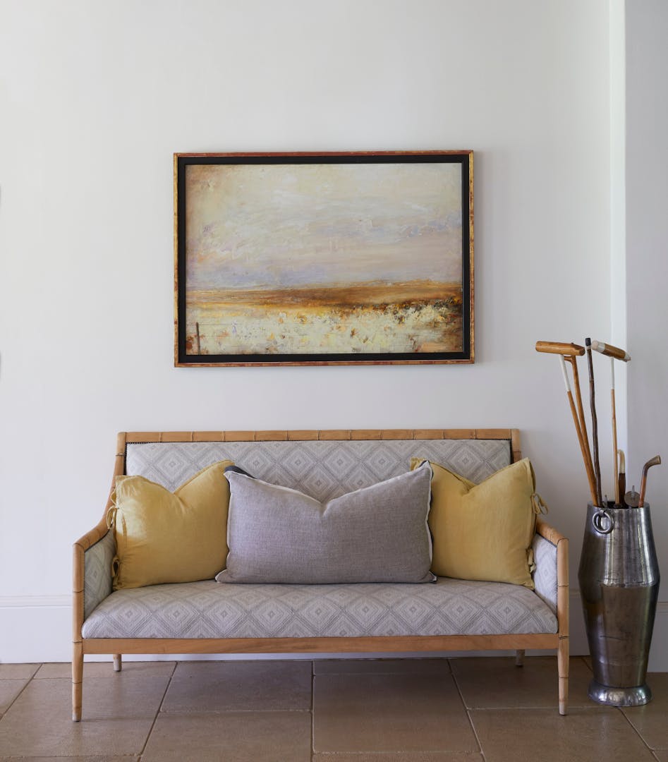 A bench upholstered in grey, geometric-patterned fabric has a light wood frame that seems to be bamboo, and holds three large cushions. It sits on a floor made of large stone tiles, with a metallic vessel beside it holding vintage sporting equipment, and a framed painting above it showing an impressionistic landscape