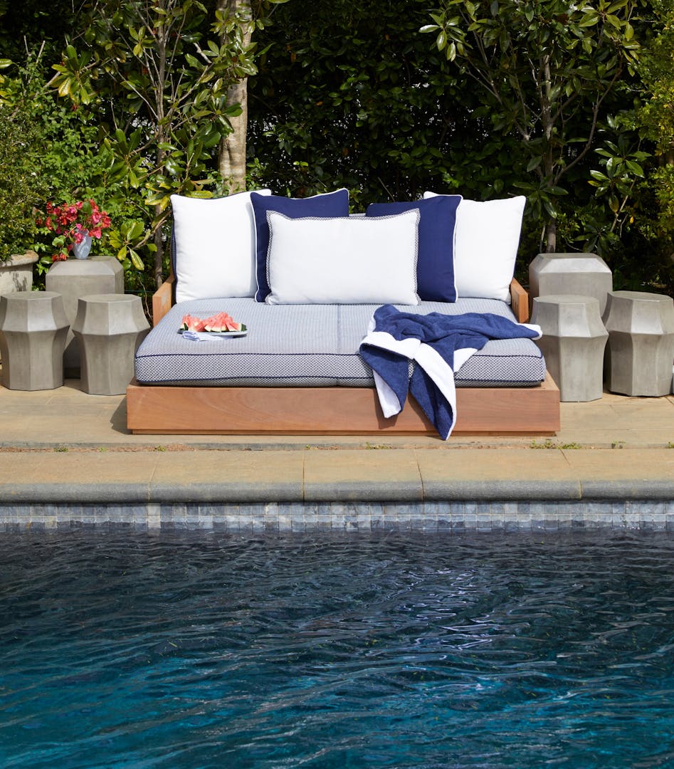 The water of a dark-bottomed swimming pool is visible in front of poolside furniture holding blue and white cushions, a blue-and-white towel, and a plate of watermelon slices. Lush greenery is in the background