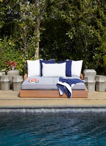 The water of a dark-bottomed swimming pool is visible in front of poolside furniture holding blue and white cushions, a blue-and-white towel, and a plate of watermelon slices. Lush greenery is in the background.