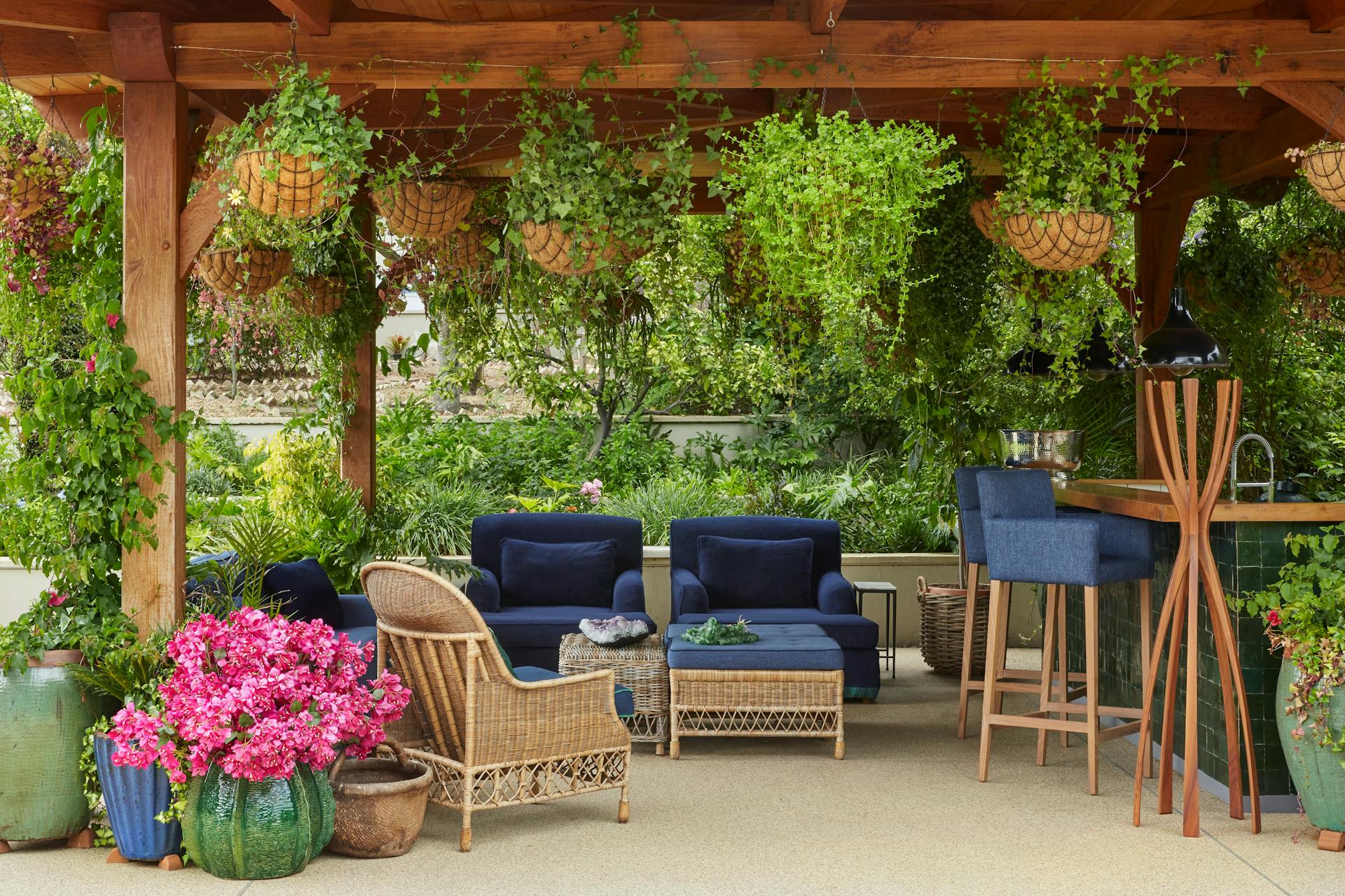 An outdoor entertaining area featuring rattan furniture, surrounded by greenery, with a bar area to the left.