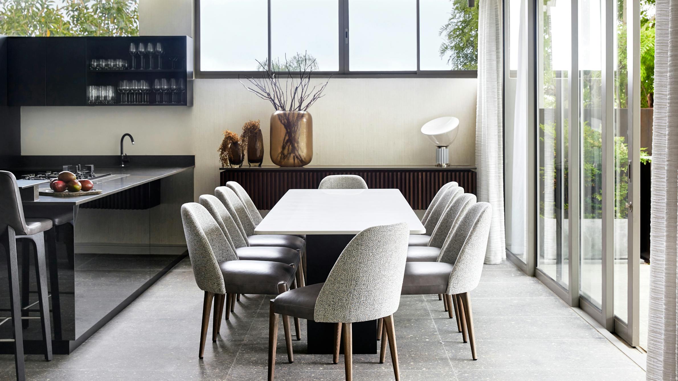 A modern dining room with grey dining chairs and a sleek dining table with sliding doors to the right and the kitchen to the left.