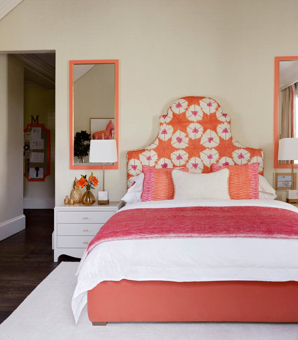 A child's bedroom with a peach-toned, patterned headboard, pink and white bedding and bedside tables on either side with mirrors hanging on the wall above them.
