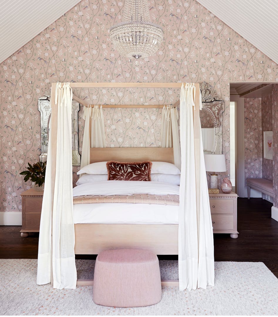 A sophisticated bedroom with a 4-poster bed in the centre, patterned wallpaper on the back wall and a pouffe just in front of the bed on the rug.